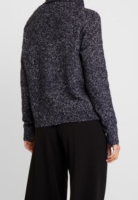 Donkerblauw pluizig trui met een hoge kraag, geribde manchetten en zoom. Bevat een textuur gebreid patroon. Gemaakt in combinatie met zwarte wijde broek.