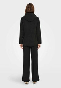 Černá bunda s kapucí, strukturovaná tkanina a upravený střih, v kombinaci s volnými černými kalhotami a špičatými podpatky.