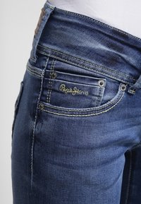 Vaqueros de mezclilla azul oscuro con un ligero efecto desgastado, con costuras doradas y un logo de "Pepe Jeans" cerca del bolsillo frontal.