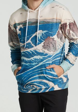 Person trägt einen blau-beigen Hoodie mit einem Design aus Ozeanwellen, Felsen und fliegenden Vögeln, Hände teilweise in der Vordertasche.