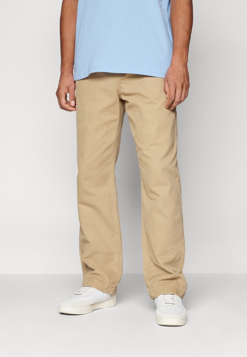 Homme portant un pantalon beige à jambes larges, une chemise bleu clair et des baskets blanches, debout devant un fond clair uni.