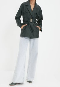 Dark green belted jacket with a collared neckline, featuring chest pockets and long sleeves, worn with light wash wide-leg jeans.