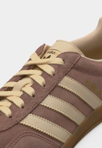 Gros plan sur une sneaker Adidas Gazelle rose et crème avec une texture en daim, des lacets crème et des lettres dorées sur le côté.