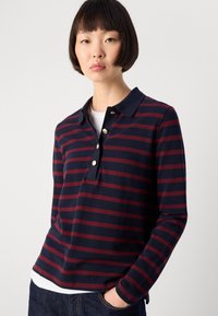 Young person with short dark hair wearing a navy and burgundy striped long-sleeve polo shirt and dark jeans, standing with one hand in pocket.