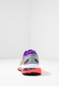 Zapatilla deportiva con una parte superior de malla gris, cuello morado, acentos naranjas y una suela blanca que presenta un clip de talón morado y una suela exterior de goma negra.