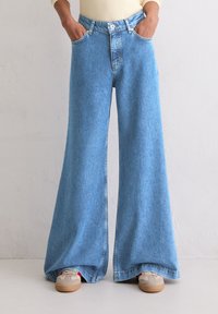 Persoon in lichtblauwe wijde broek en beige sneakers met handen in de zakken tegen een effen wand achtergrond.