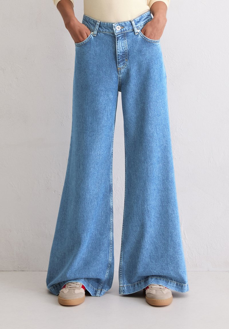 Persoon in lichtblauwe wijde broek en beige sneakers met handen in de zakken tegen een effen wand achtergrond.