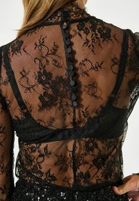 Sort blonderet top med intrikate blomster mønstre, transparent mesh og en række cirkulære sorte knapper på ryggen. Ærmerne er gennemsigtige.