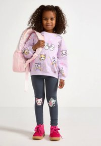 Fialová mikina s kreslenými mačkami, sivé legíny s nášivkami mačiek na kolenách a ružové plyšové členkové topánky. Ružový batoh s sieťovinovými detailmi.