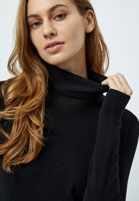 Maglione nero a collo alto con colletto e polsini a costine, realizzato in un morbido tessuto testurizzato. Primo piano sulla scollatura e sull'area superiore del torace.