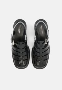 Ζευγάρι μαύρα δερμάτινα σανδάλια ψαράδων με λουριά που κλείνουν με ασημένιες αγκράφες και υποδήματα με λογότυπο, τοποθετημένα δίπλα δίπλα σε λευκό φόντο.