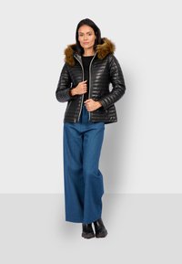 Černá prošívaná bunda s kapucí lemovanou kožešinou, zapínáním na zip vpředu a bočními kapsami, doplněná širokými modrými kalhotami a černými botami.