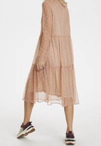 Lång, lätt, genomskinlig klänning i en mjuk persikofärg med blommönster, flerlagersdesign och långa ärmar. Kombinerad med sportiga sneakers.