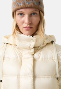 Kurtka puchowa w kremowym kolorze z wysokim kołnierzem, wyposażona w zapięcia na zatrzaski i sznurki do regulacji. W komplecie dziergany czapka w brązowo-szary wzór.