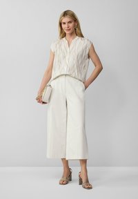 Weiße, strukturierte ärmellose Bluse mit Wellenmuster, kombiniert mit schimmernden, weiten Hosen. Accessoires sind eine kleine Clutch und Riemchensandalen.