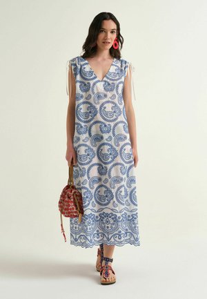 Donna che indossa un vestito lungo bianco con motivi paisley blu, orecchini rossi, borsa rossa a fantasia e sandali rossi e blu abbinati.