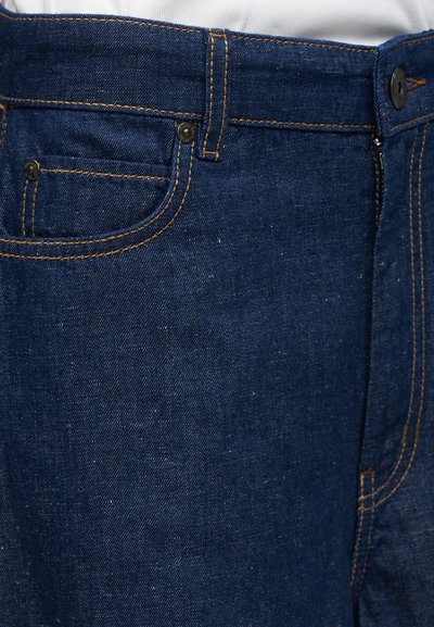 Σκούρο μπλε τζιν μπροστά με ορατή τσέπη, μεταλλικά καρφιά, ραμμένα ραφές και μαύρο κουμπί στη μέση.