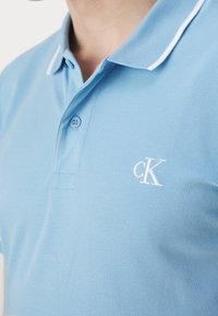 Jasnoniebieska koszulka polo z teksturowanej tkaniny, z białym wykończeniem kołnierzyka i małym białym logo "CK" na piersi.