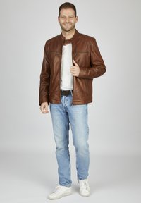 Braune Lederjacke mit Stehkragen, zwei Reißverschluss-Taschen und gesteppten Schulterakzenten. Getragen über einem grauen Hemd und blauen Jeans.