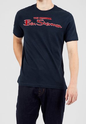 Mężczyzna w granatowym t-shircie z krótkim rękawem z czerwonym napisem „The Original Ben Sherman” na piersi i ciemnoniebieskich dżinsach.