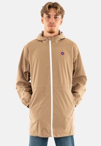 Imperméable beige léger avec capuche, doté d'une fermeture éclair blanche et d'un petit logo circulaire rouge et bleu sur la poitrine. Texture lisse.