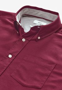 Camicia button-up bordeaux con colletto strutturato, caratterizzata da una fodera a righe orizzontali, bottoni bianchi e una tasca sul petto.