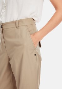 Beige Hosen mit einer glatten Textur, Seitentaschen und einem dekorativen Logo-Akzent auf der linken Seite. Der Bund ist flach mit einem einfachen Knopf.