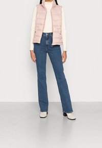 Chaleco acolchado rosa claro con cuello alto, combinado con jeans ajustados azul y botas blancas hasta el tobillo, creando un conjunto casual en capas.