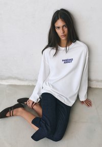 Jonge vrouw met lang donker haar zittend op de vloer, draagt een wit shirt met lange mouwen met de tekst "PANCAKE ADDICT" en zwarte broek en schoenen.