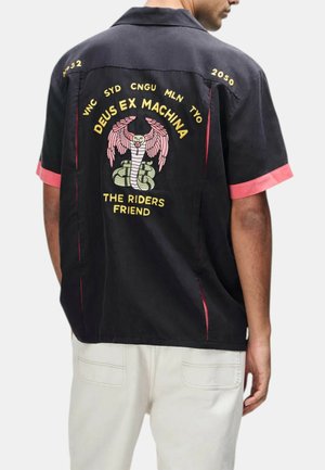 Uomo con camicia nera a maniche corte con polsini rosa, ricamata con cobra e ali e testo "DEUS EX MACHINA THE RIDERS FRIEND" sulla schiena.