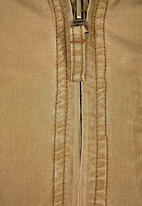 Beige tyg med en slät yta med en central dragkedja. Dragkedjehandtaget visar märket "SCHOTT." Väl sydda kanter fulländar designen.