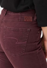 Burgunderfarbene Denim-Jeans verfügen über eine Rückentasche mit pinkfarbener Naht und einem gebrandeten Lederpatch mit Sternenmuster am Bund.