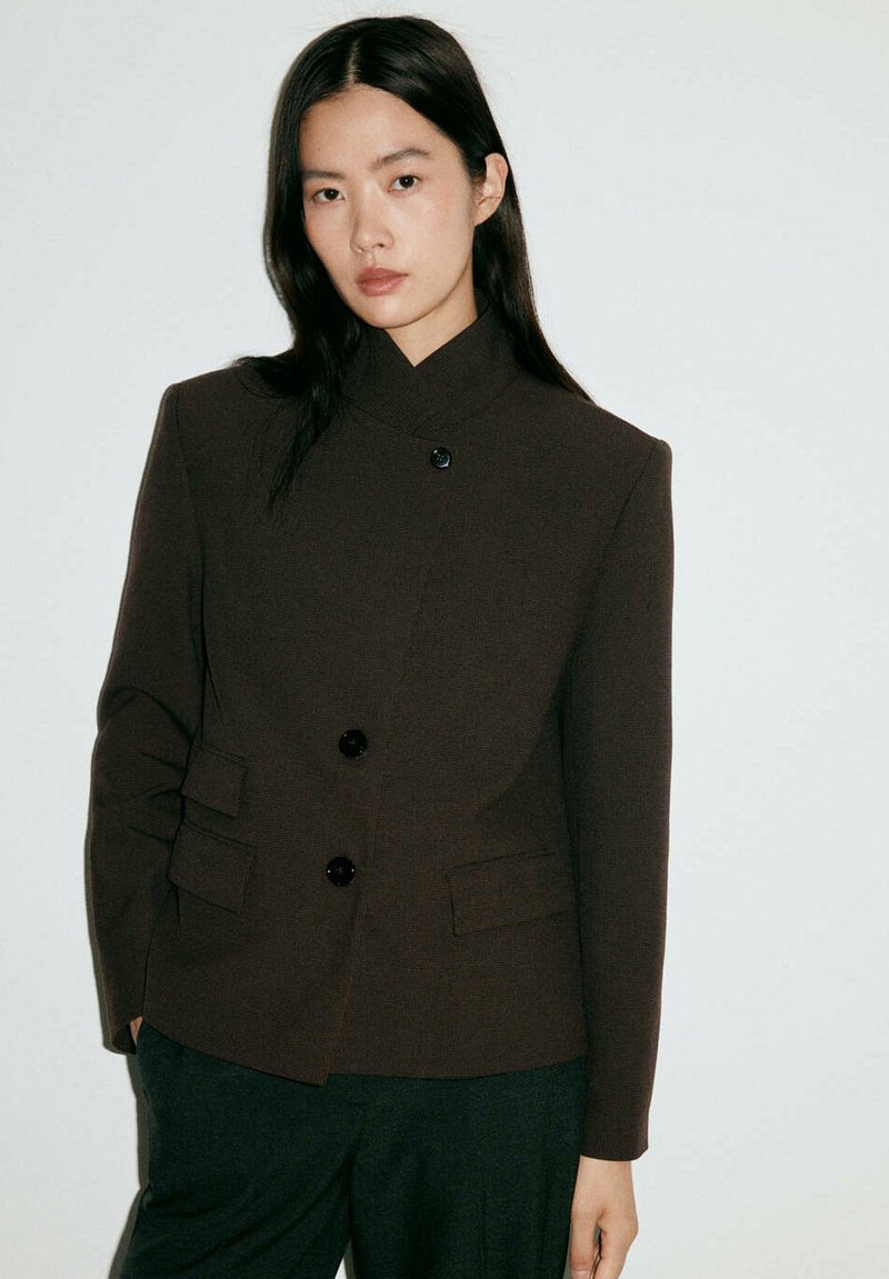 Jeune femme aux longs cheveux foncés portant un blazer marron foncé boutonné et un pantalon sombre sur un fond clair uni.