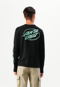 Man in een zwart langemouwen shirt met een mintgroene "Santa Cruz Skateboards" logo op de rug en de linkermouw, gecombineerd met beige cargo shorts.