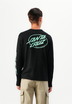 Man in een zwart langemouwen shirt met een mintgroene "Santa Cruz Skateboards" logo op de rug en de linkermouw, gecombineerd met beige cargo shorts.