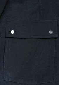 Mörk marinblått tyg med en stor ficka med klaff och silverknapp. Ytan verkar slät med synliga söm detaljer.