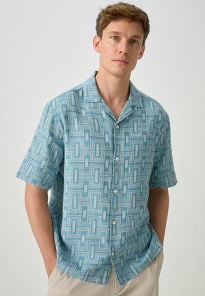 Junger Mann trägt ein hellblaues, kurzärmeliges Hemd mit Knöpfen und geometrischem Muster sowie eine beige Hose, steht vor einem schlichten Hintergrund.