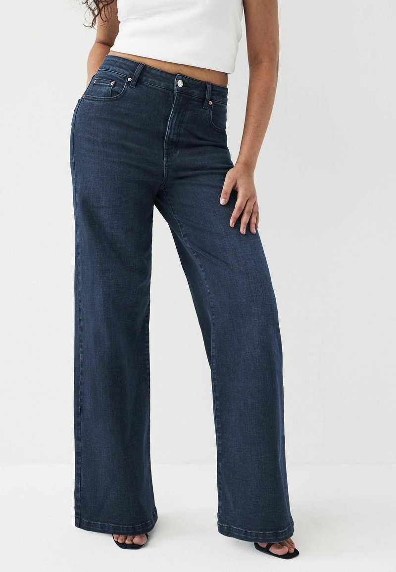 Femme portant un jean bleu foncé taille haute à jambes larges, associé à un top court blanc sans manches et des talons ouverts noirs.