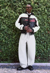 Giacca varsity bianca e nera con dettagli rossi, abbinata a pantaloni sportivi grigi chiari a gamba larga e scarpe nere lucide. Gli occhiali da sole completano il look.