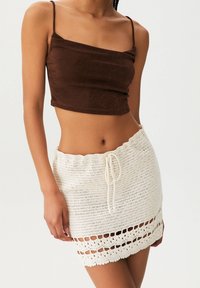 Vrouw in een bruin cropped tanktop en een witte gehaakte minirok, gestrikt op de taille, tegen een effen achtergrond.