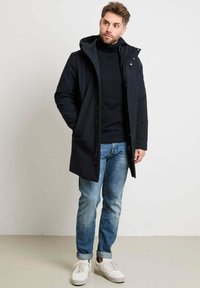 Zwarte parka jas met een capuchon, ritssluiting en drukknoopsluiting, gedragen over een donkere coltrui, gecombineerd met lichtblauwe jeans en witte sneakers.