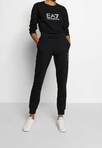 Svart sweatshirt- och joggers-set tillverkat av mjukt material. Innehåller en logotyp på bröstet och en dragsko i midjan. Fulländat med vita sneakers.