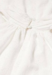 Robe en tissu blanc avec des motifs floraux texturés, dotée de manches bouffantes et de deux boutons ronds à la taille. Détail de jupe plissée.