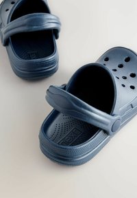 Marinblå plastklädda träskor med ventilationshål, bakkäpp, texturerade innersulor och cirkulärt mönster på sulorna.