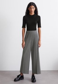 Schwarzes, geripptes T-Shirt mit kurzen Ärmeln, hochgeschnittene graue Weitbeinhose mit vertikalen Streifen und schwarze Schnürschuhe mit dicker Sohle. Glatte Stoffstruktur.