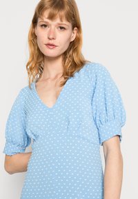 Vestido azul claro com mangas curtas e volumosas. Apresenta um decote em V profundo e um padrão de bolinhas brancas. Textura de tecido suave.