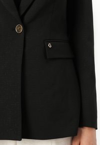 Blazer nero in tessuto testurizzato, chiusura con un bottone e tasca decorata con un logo metallico. Vestibilità tailored, design classico.