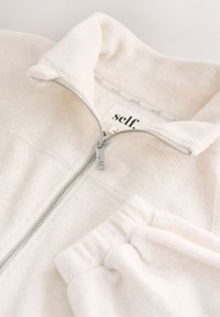 Veste polaire douce blanc cassé avec fermeture éclair et pantalon polaire assorti, tous deux affichant les étiquettes de la marque « self » et une tirette de fermeture éclair en métal.