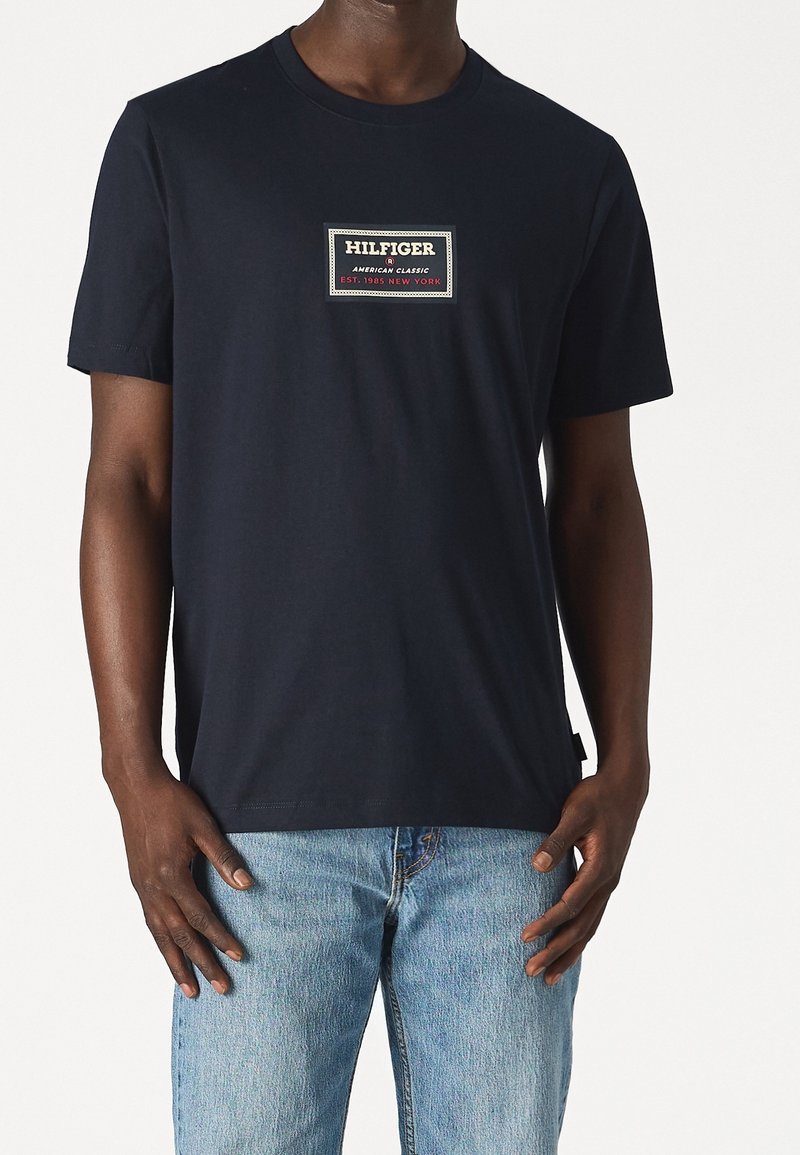 Camiseta de algodón azul marino con un parche rectangular de logo que dice "HILFIGER" en blanco, rojo y verde. Mangas cortas, cuello redondo.