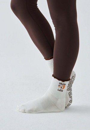 Nogi w brązowych legginsach noszą białe antypoślizgowe skarpetki z napisem „Pilates & Latte” i wzorem filiżanki kawy, podeszwy ozdobione uchwytami w kształcie serc.